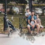 Vader zit met drie kinderen op bankje in park naast kinderwagen